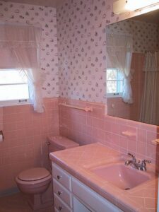 A 1960s pink bathroom with floral wallpaper, featuring a matching pink sink, bathtub, and toilet, complemented by white cabinetry and sheer window curtains.