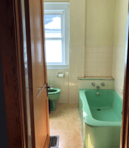 View from a hallway into a small bathroom with a green toilet and bathtub, beige floor, and white tiled walls, illuminated by natural light from a window.