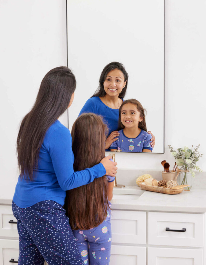 A mother and daughter in pajamas standing in front of a mirror.